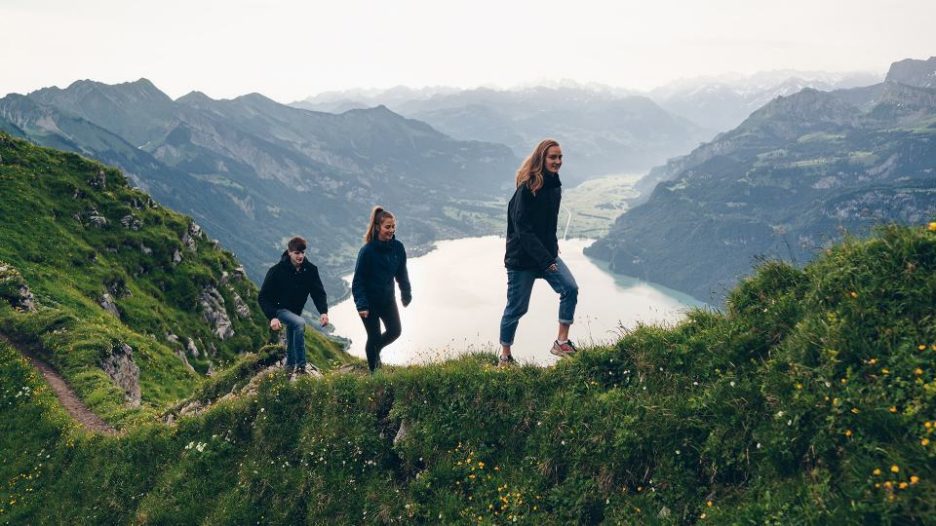 group of two females and a male hiking up a hillside on the edge of a lake with mountains in the distance