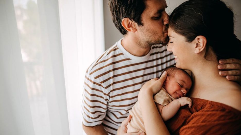 a man kisses his wife on the forehead while she holds a young sleeping baby