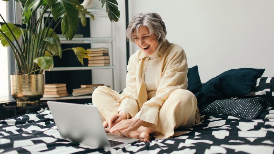 Happy mature woman using notebook while working remotely on project from home and sitting on bed