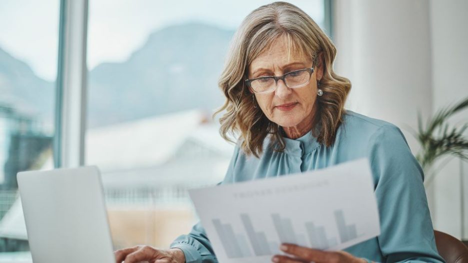 a ssenior manager with glasses is sitting at her desk near a window and reading a financial report while using her laptop