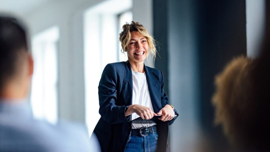 Beautiful cheerful woman in a dark blue jacket standing in front of a crowd looking down at a tablet.