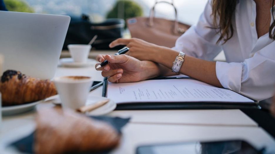 close up of a woman's hands holding a pen with a laptop, clipboard, coffees and pastries nearby