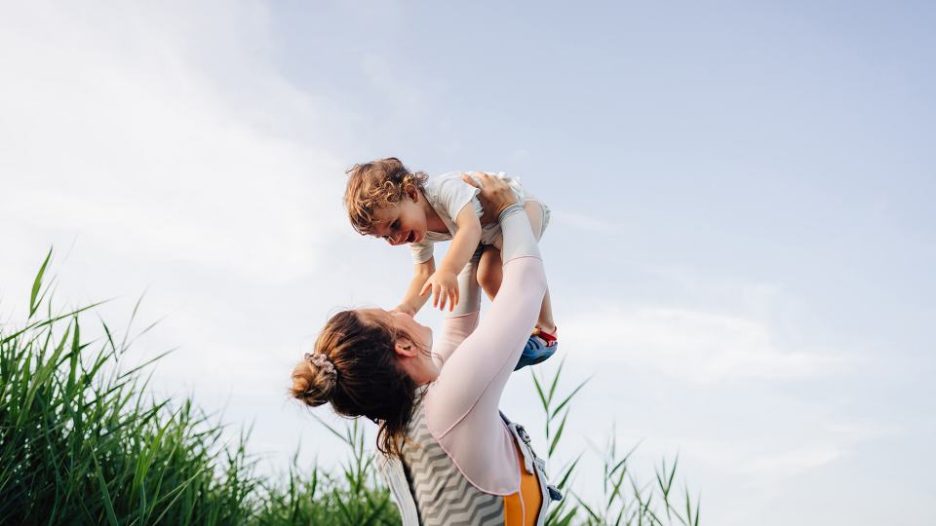 a mother lists her toddler overhead while they're outside in a grassy field