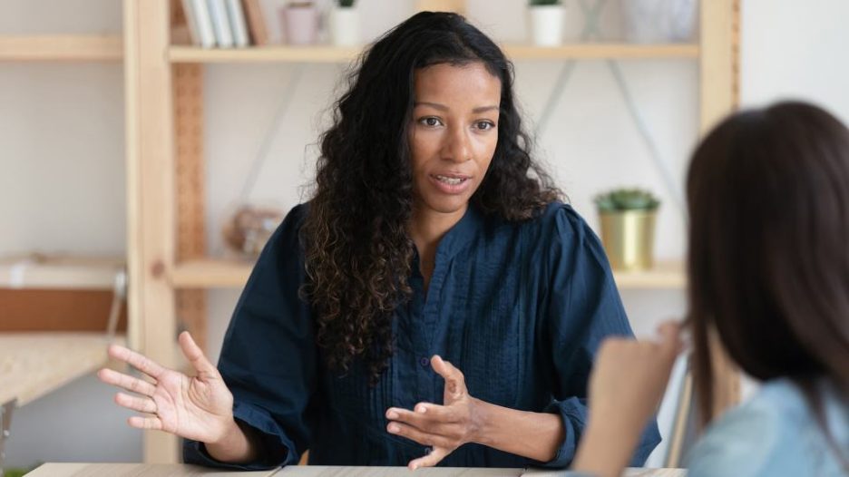 two women are meeting in a modern office with bookcases and one woman is explaining something