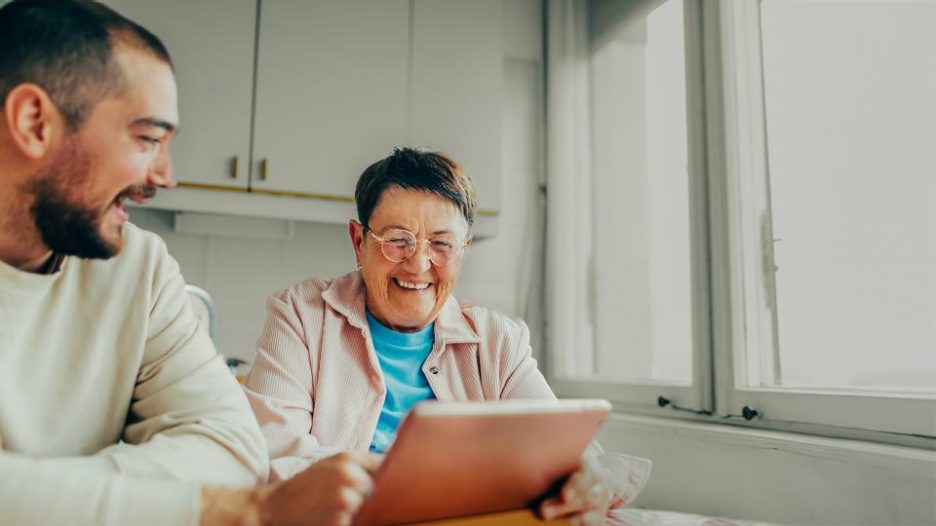 mature woman smiles while she looks at her tablet and a young man is sitting beside her also smiling