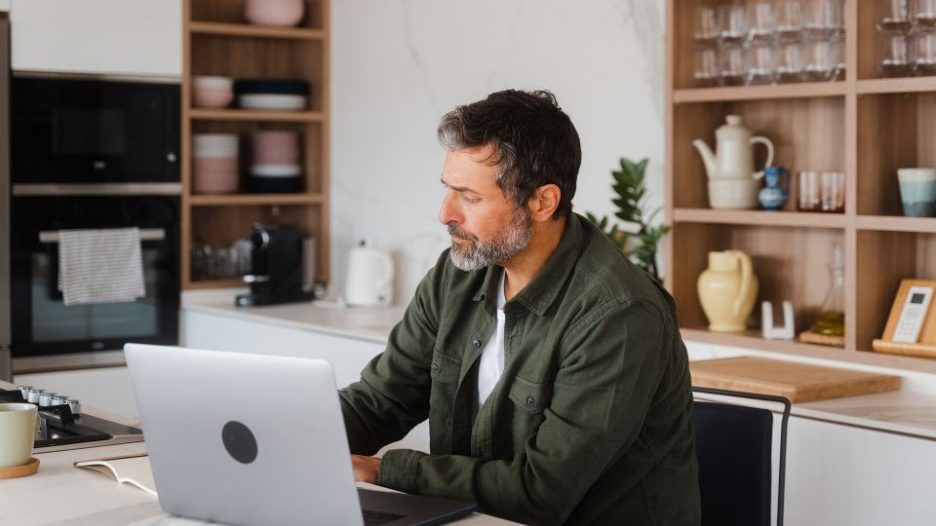 man in a striped shirt using his laptop and taking notes in a home office