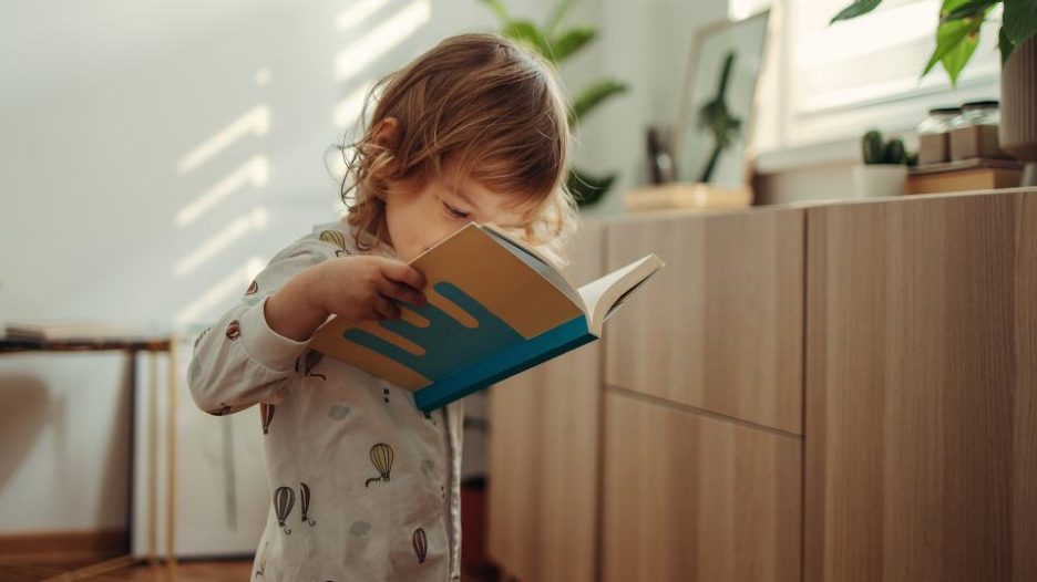 young child in pyjamas looking closely at a book in her room